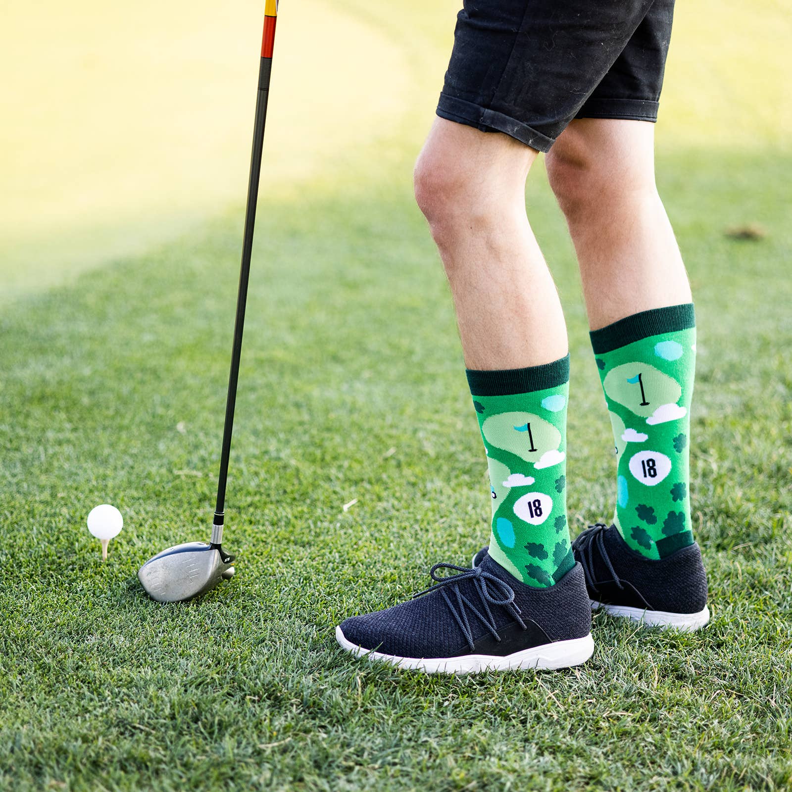 man's legs at a golf course with an 9 iron in front of a golf ball on a tee wearing black shorts, black shoes and green golf socks