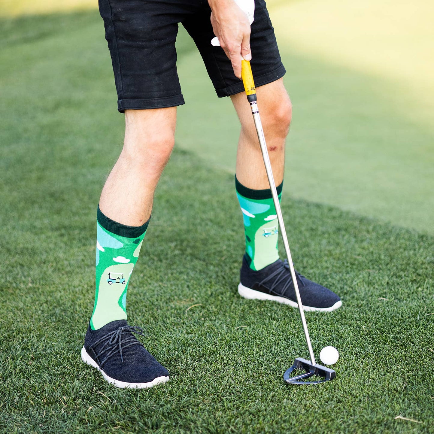 man's legs at a golf course with a putting iron in front of a golf ball  wearing black shorts, black shoes and green golf socks 