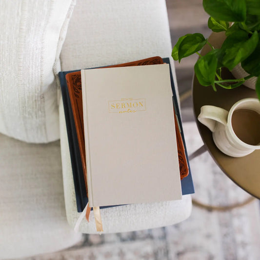 white hard cover book with gold foil writing that reads "Sermon Notes" laying on topo f a brown journal and dark blue bible on the arm of a white couch, coffee cup on side table