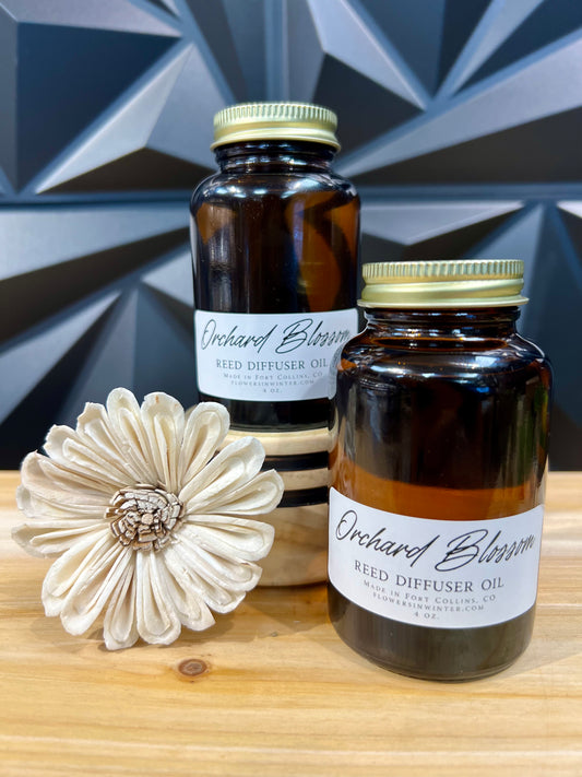 two mason jars with gold lids, white lables that reads Orchard Blossom Reed Diffuser Oil on a wood table, textured black wall in the back and a flower reed diffuser on the table