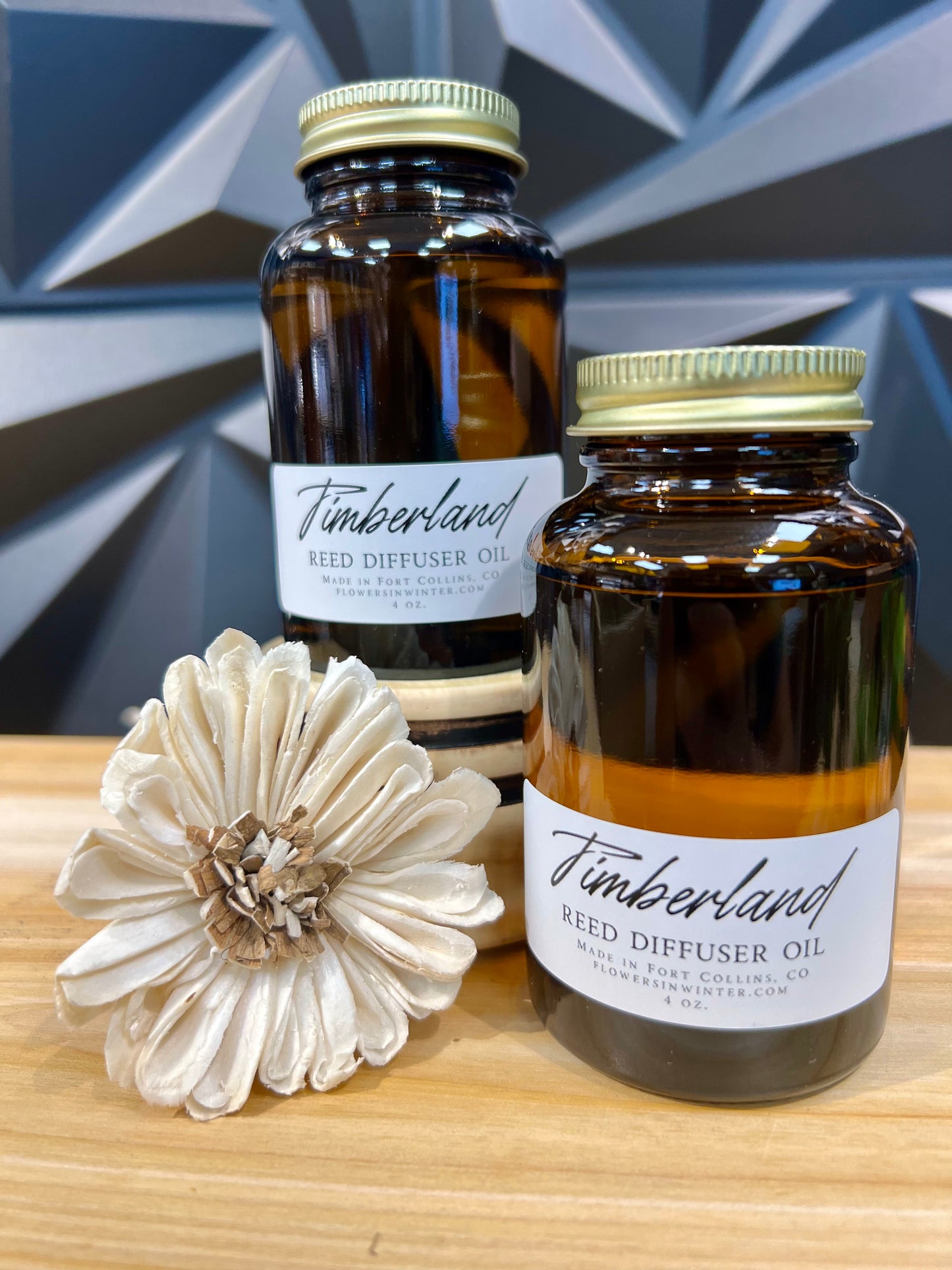 two mason jars with gold lids, labels are white with black letters that read "Timerland Reed Diffuser Oil" on a table next to flower reed