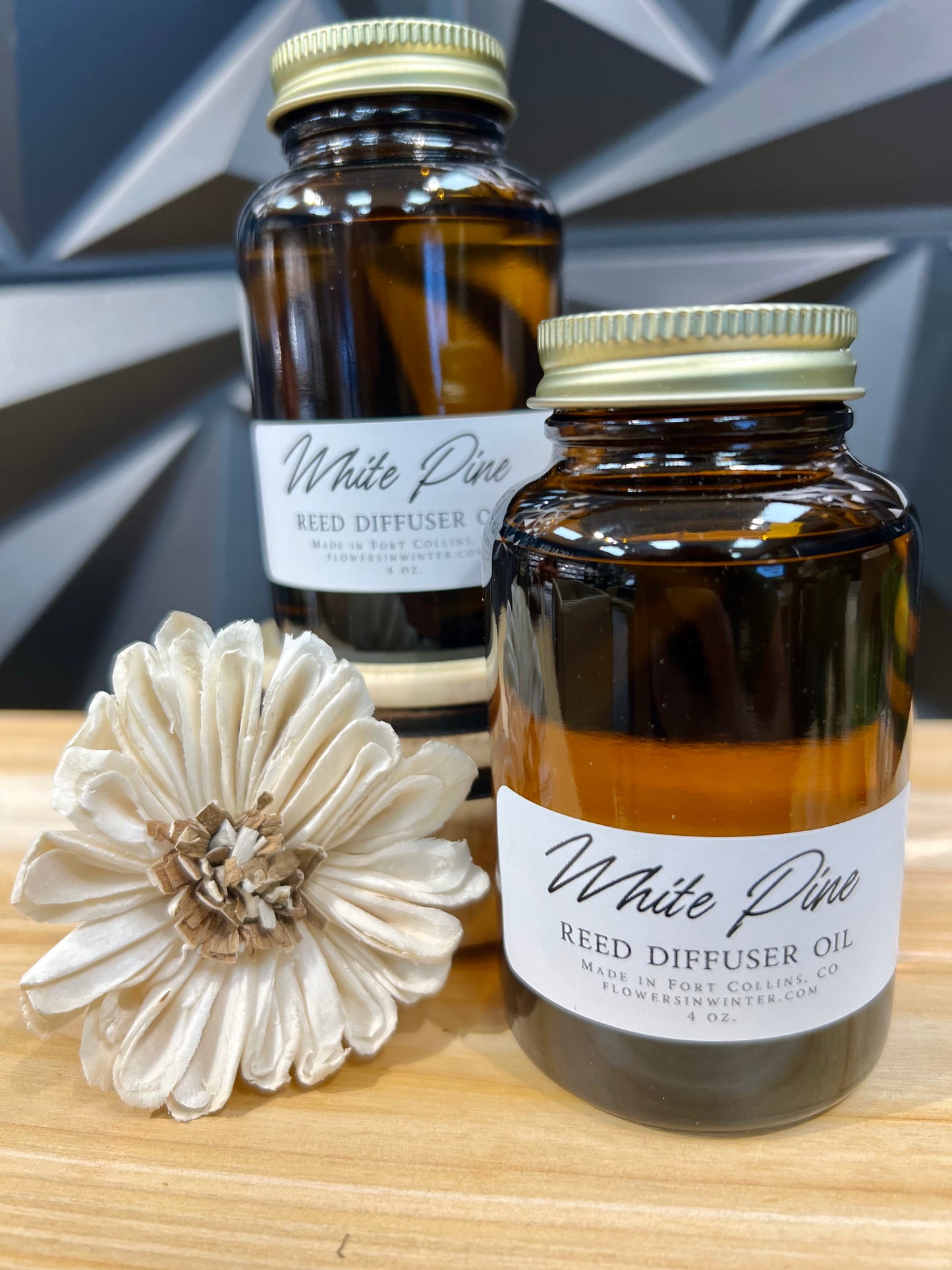 two mason jars with gold lids, white labels read "White Pine Reed Diffuser Oil" sitting on wood table next to flower reed