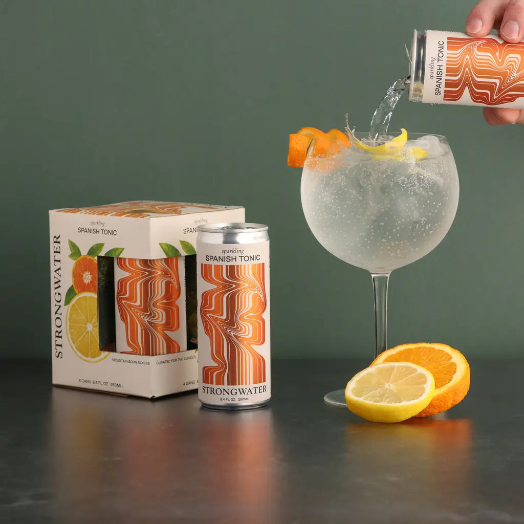 pack of four Sparkling Spanic Tonic, one can out of the box, a person pouring another can into a large clear margarita style galss with orange and lemon slices