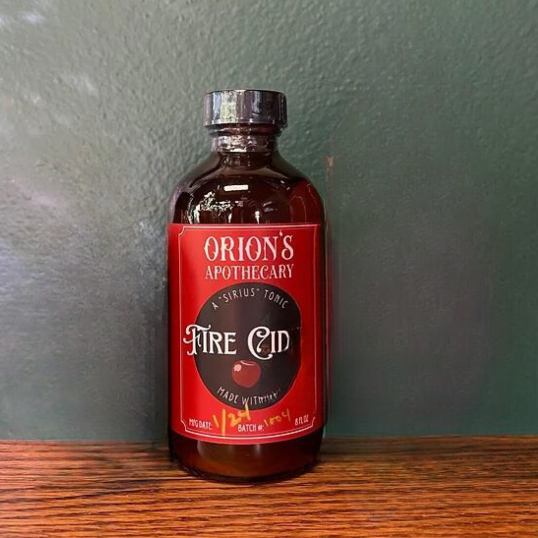 dark amber glass bottle with black lid, and red label on table that reads " Orion's Apothecary Fire Cider"