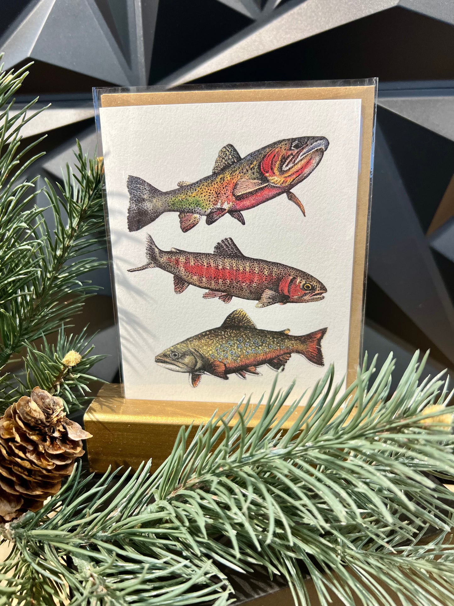 greeting card with three trout, on top of a tan envelop sitting on a table with pine branch