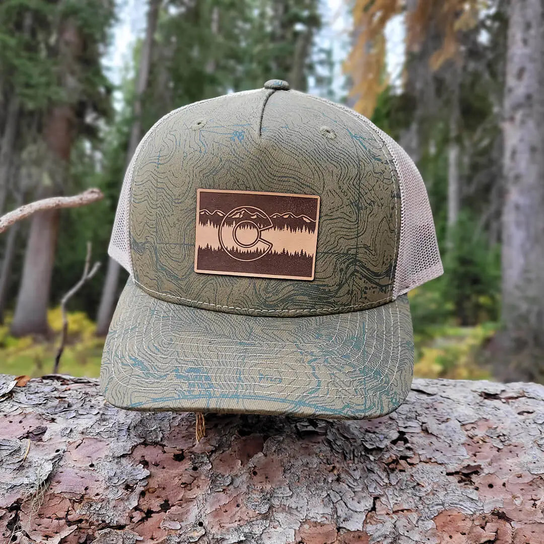 green trucker hat with topographical map of Colorado and a brown Colorado C patch outside on a tree branch