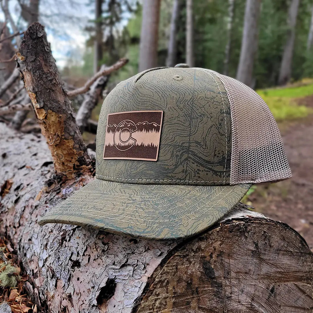 side view of green trucker hat with topographical map of Colorado and a brown Colorado C patch on a log