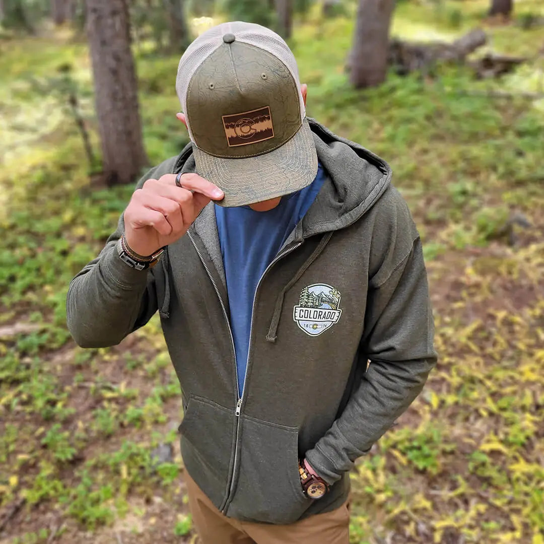 man wearing green trucker hat with topographical map of Colorado and a brown Colorado C patch outside in the woods