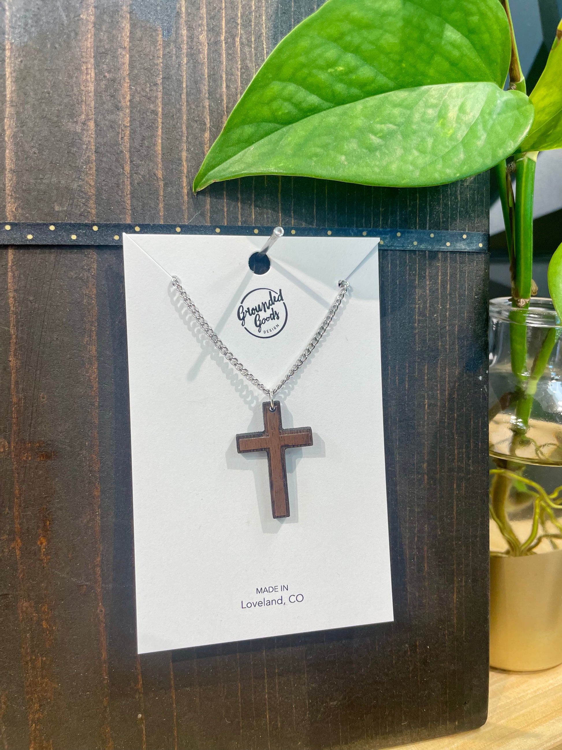 two toned wood cross on silver chain displayed on white hanging card that states "Made in Loveland, CO" leaning on a brown board on a wood table