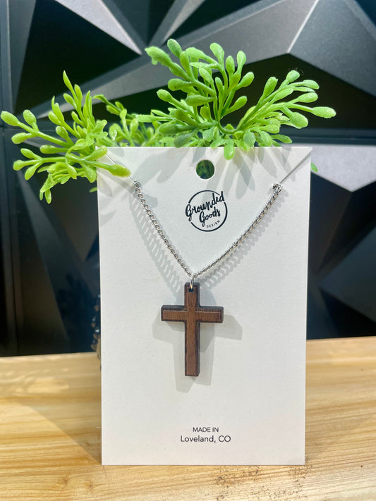 two toned wood cross on silver chain displayed on white hanging card that states "Made in Loveland, CO" leaning on a small green plan on a wood table