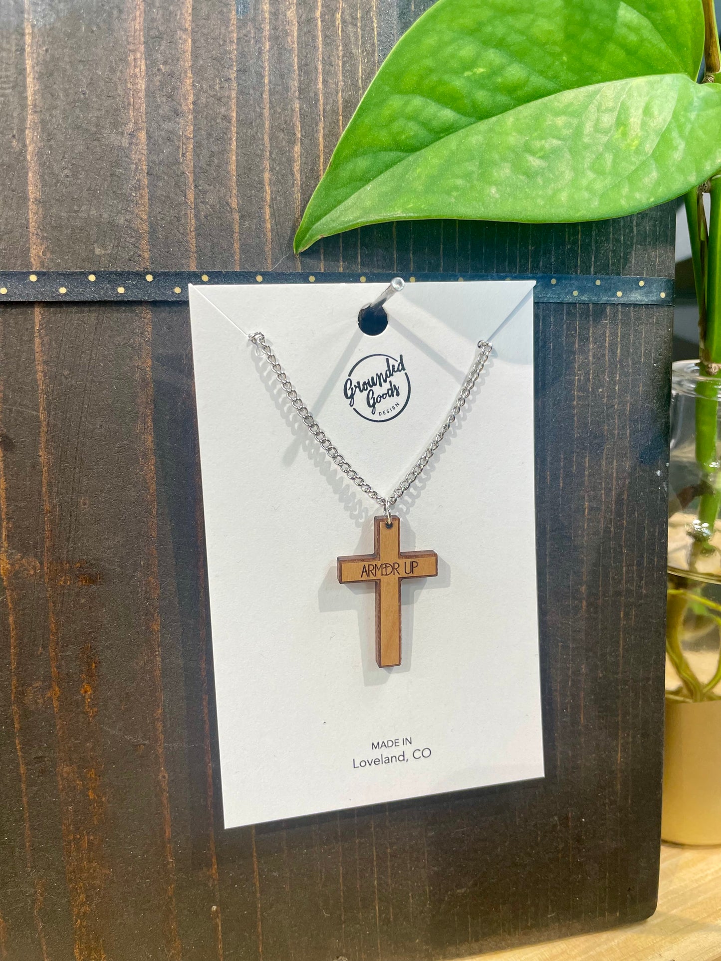 wood cross with the words "ARMED UP" etched across the arms on a white display card sitting on a wood table in front of a green plant slightly tilted to the right