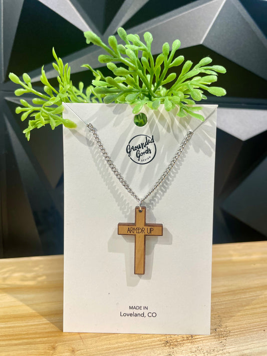 wood cross with the words "ARMED UP" etched across the arms on a white display card sitting on a wood table in front of a green plant