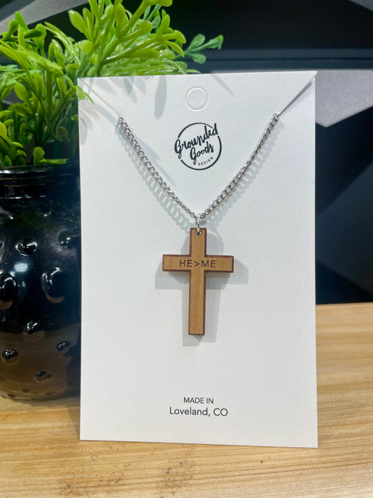 wood cross necklace etched with "He>Me" (meaning He is Greater than Me) across the arms on a silver chain on a white display card sitting next to a green plan on a wood table