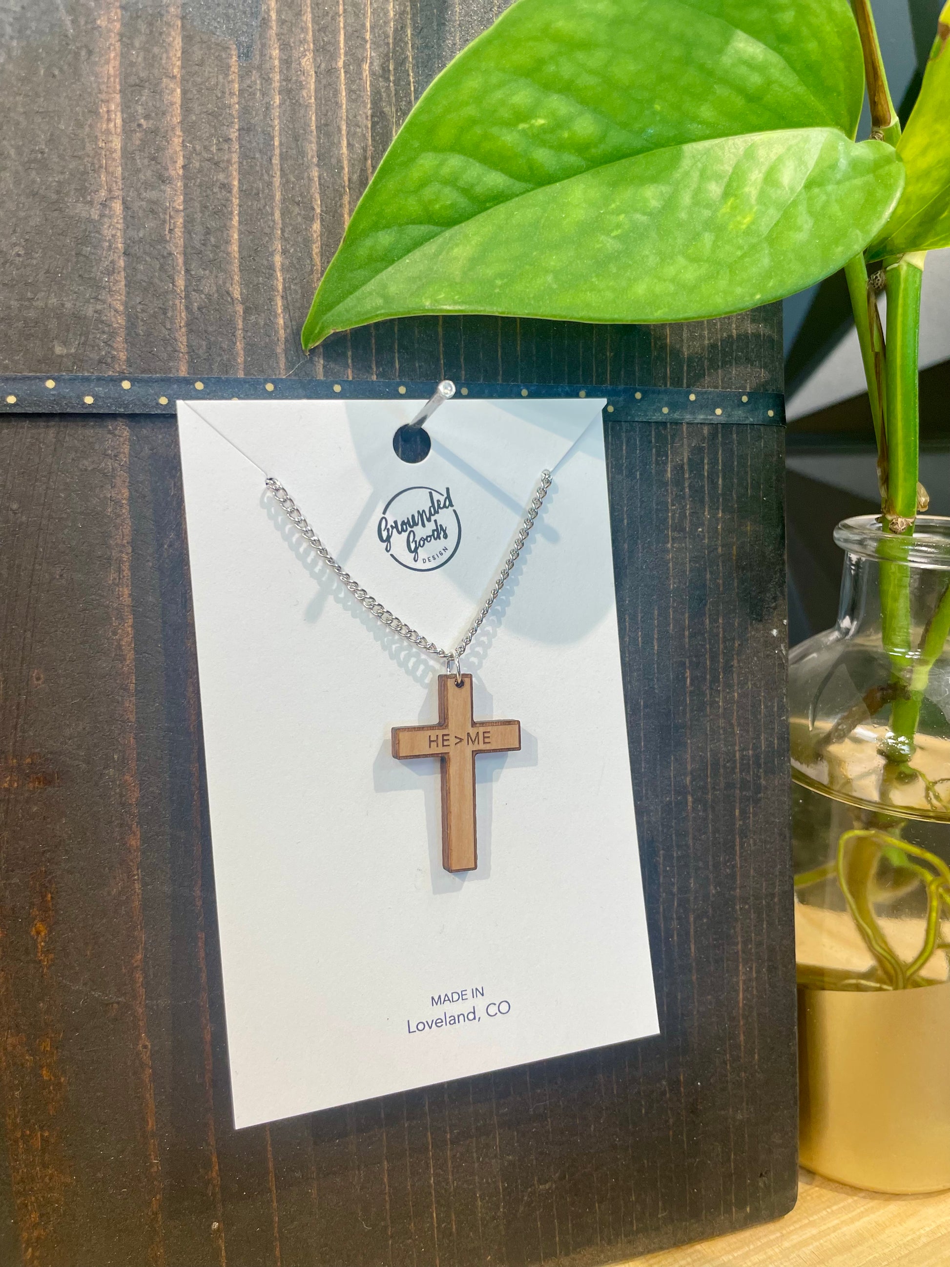 wood cross necklace etched with "He>Me" (meaning He is Greater than Me) across the arms on a silver chain on a white display card sitting next to a green plan on a wood table