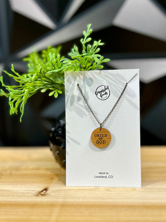 round wood pendant that reads "Child of God" on brass chain displayed on white hanging card in front of a green plant on a wood table