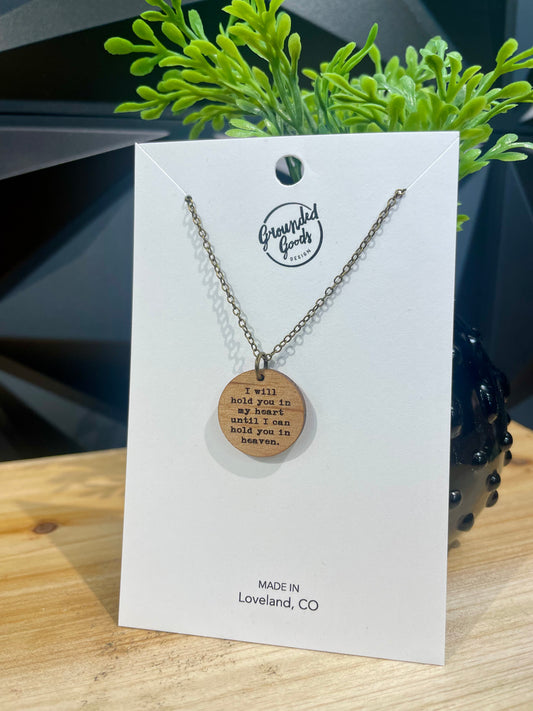 round wood pendant on a brass chain that reads "I will hold you in my heart until I can hold you in heaven" on a white display card in front of a green plant on a wood table