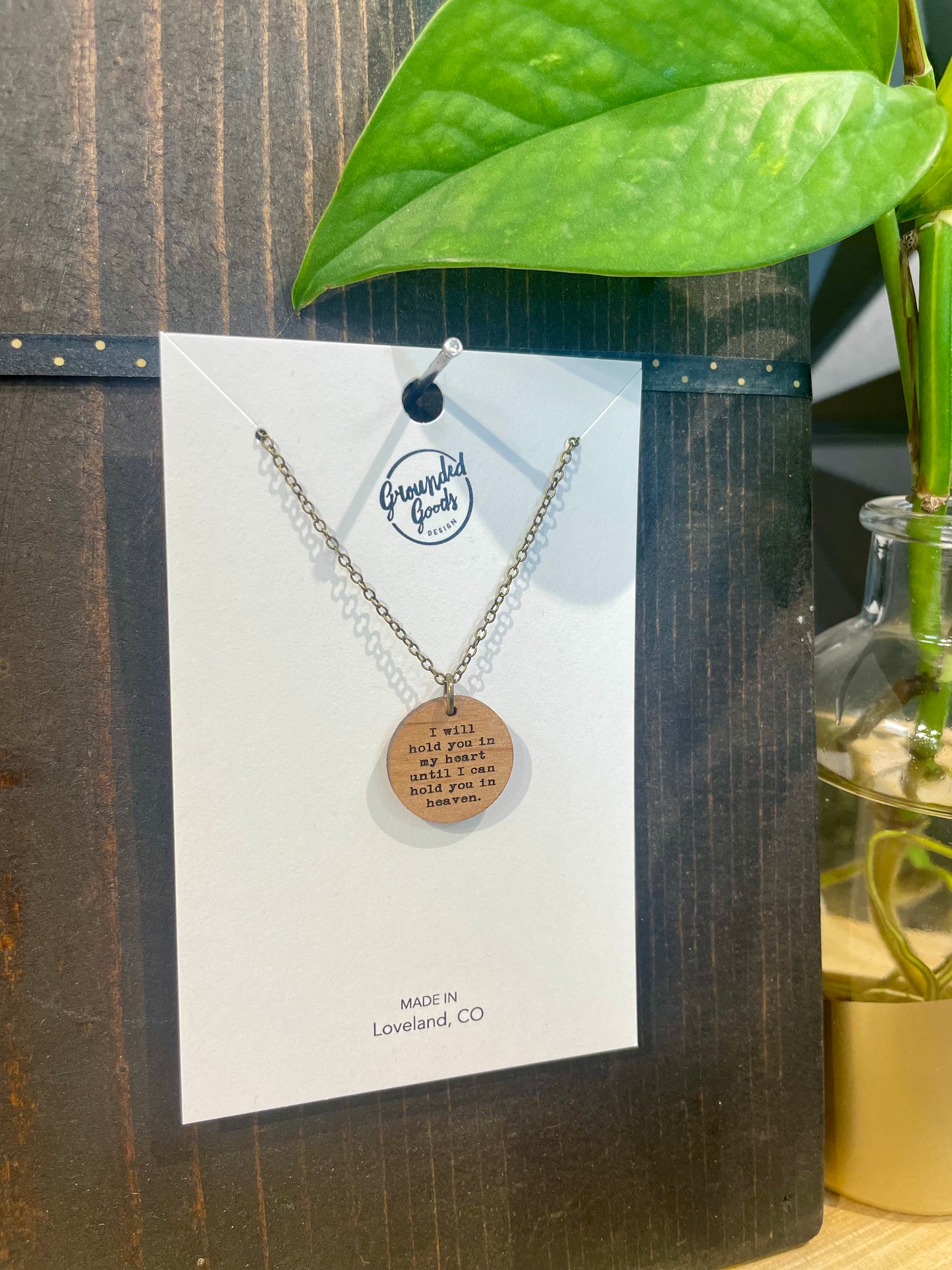 round wood pendant on a brass chain that reads "I will hold you in my heart until I can hold you in heaven" on a white display card in front of a green plant on a wood table