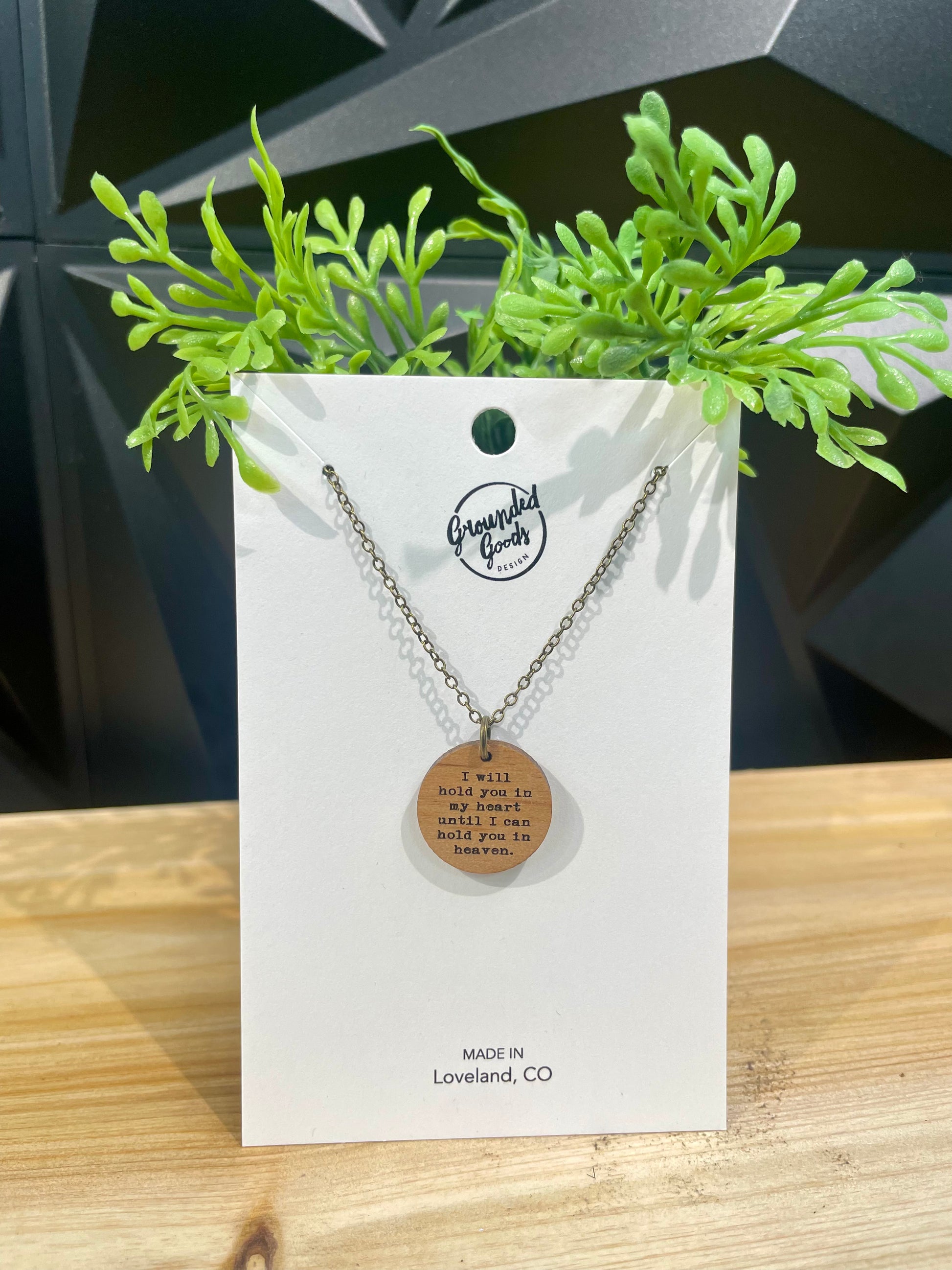 round wood pendant on a brass chain that reads "I will hold you in my heart until I can hold you in heaven" on a white display card in front of a green plant on a wood table