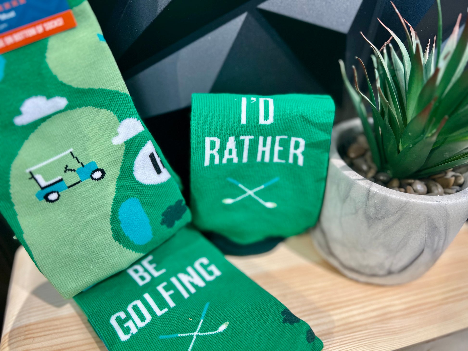 green socks displayed next to plant on wood platform, socks have gold scene and read "I'd Rather Be Golfing"