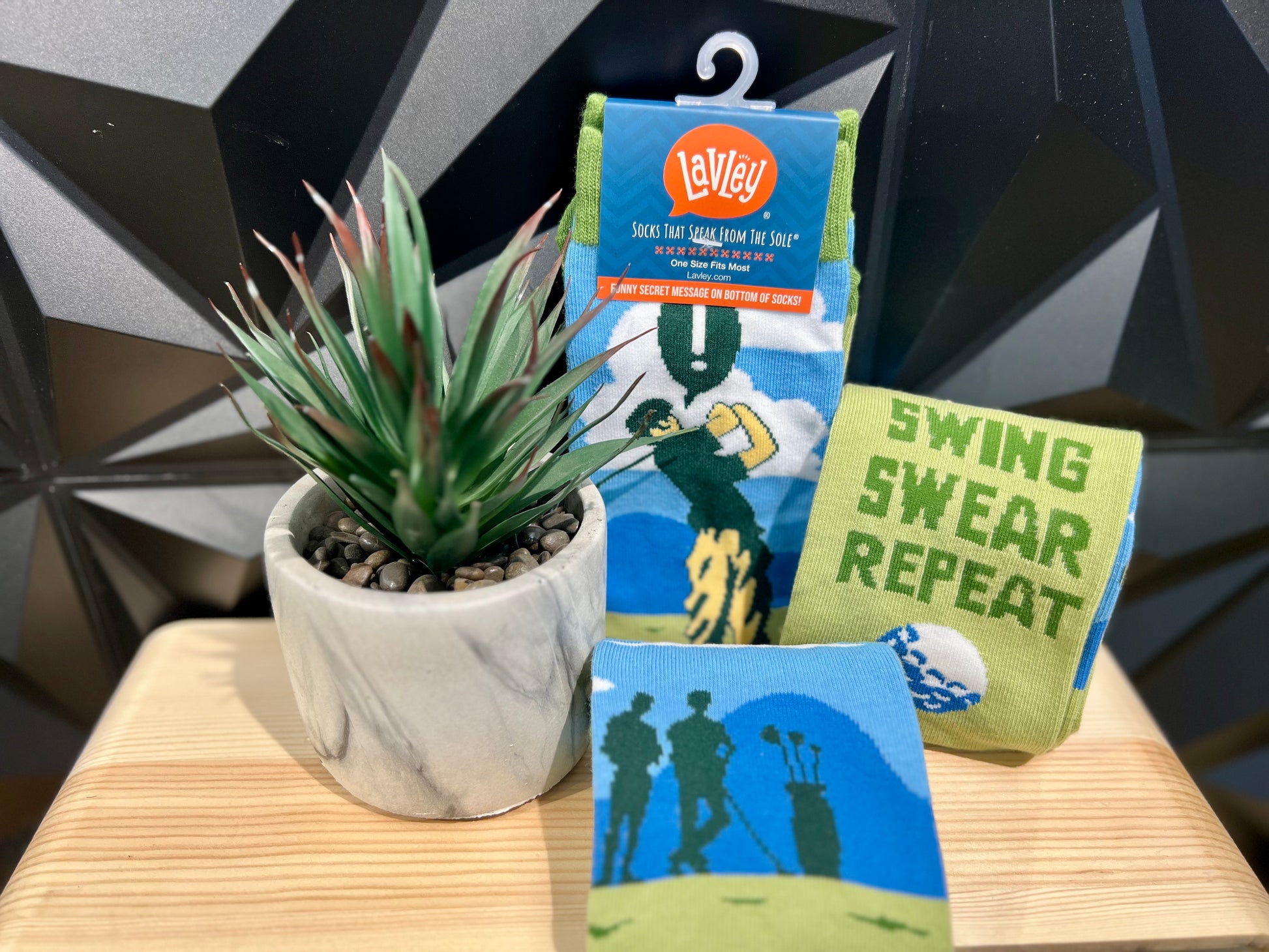 green and blue golf theme socks that read "Swing Swear Repeat" on wood stand by plant