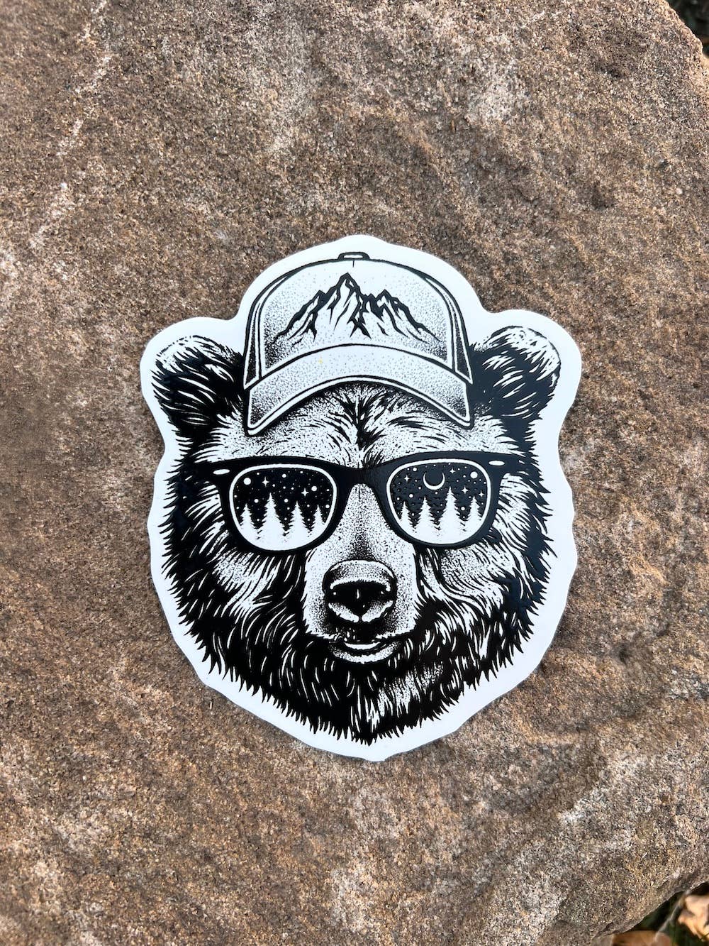 grizzle bear head sticker in black and white, he is wearing a ball cap with mountains on the front and sunglasses with trees on the lenses