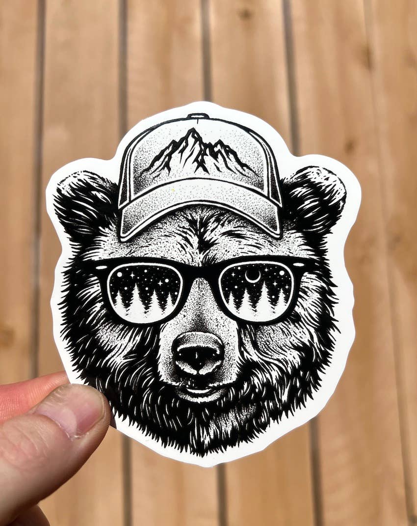 tip of persons fingers holding black and white grizzle bear head sticker, bear is wearing a ball cap with mountains on the front and sunglasses with trees in the lenses