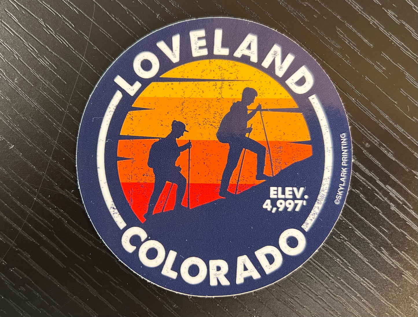 round dark blue sticker - outter edge in white says LOVELAND COLORADO Elev. 4,997 - picture in the middle is the shadow of two people hiking up a hill with a sunset in the background