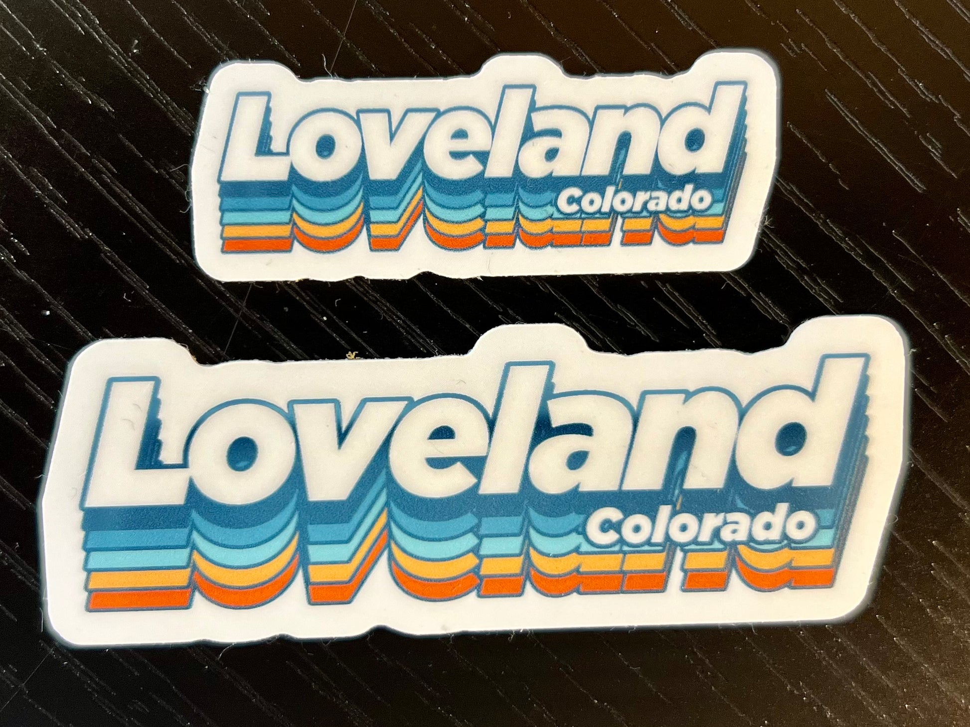 two sizes of stickers, die cut around the word LOVELAND in a retro font white, blue, light blue, yellow and orange