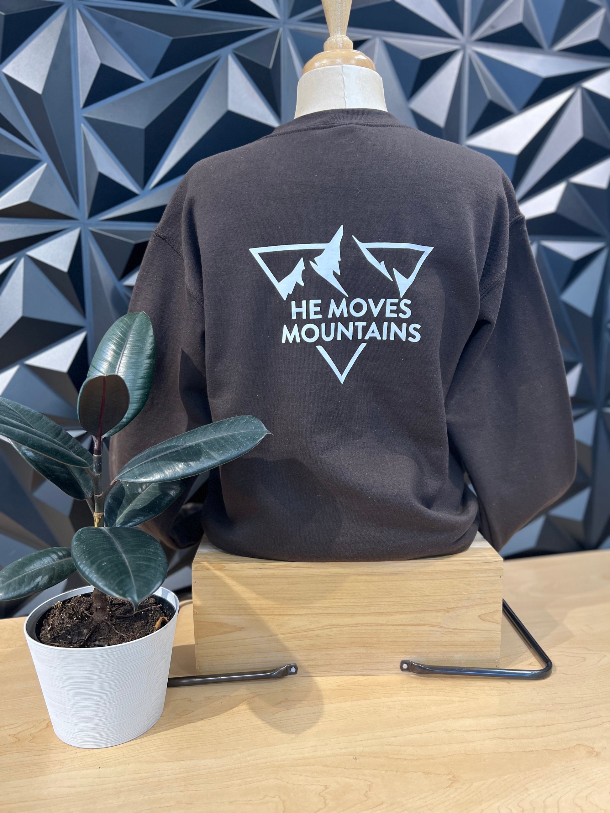 mannequin wearing a dark blue crewneck sweatshirt, showing the back that has a triangle with mountains peaks words saying HE MOVES MOUNTAINS