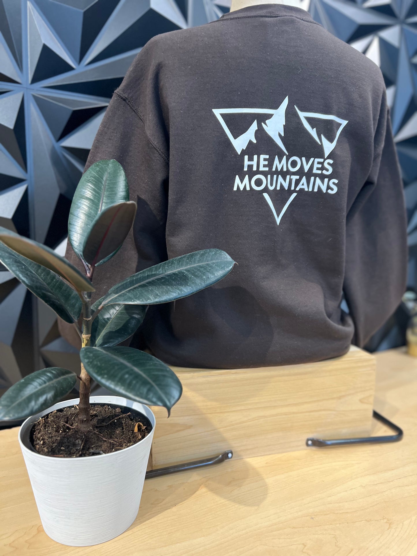 mannequin wearing a dark blue crewneck sweatshirt, showing the back that has a triangle with mountains peaks words saying HE MOVES MOUNTAINS